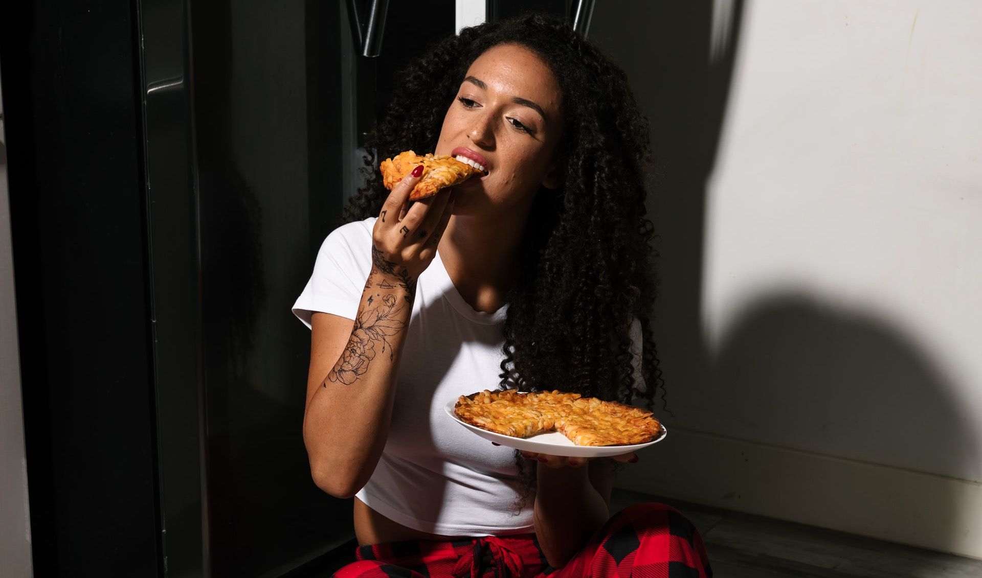 person sits on kitchen floor next to fridge, eating a pizza late at night