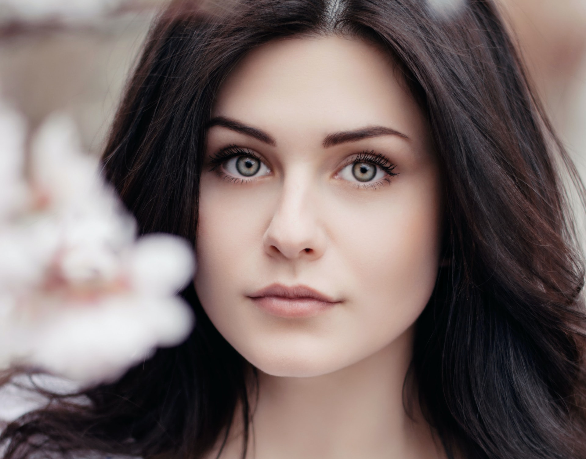 closeup of pretty person with clear skin and dark hair, looking at camera, flower to the side