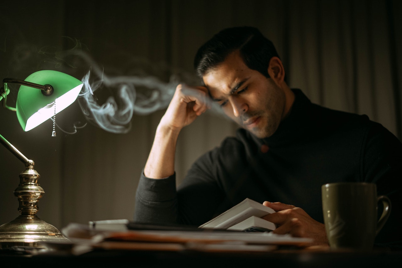 man sits at desk, head supported by fist, reading, hot cup of coffee to the side
