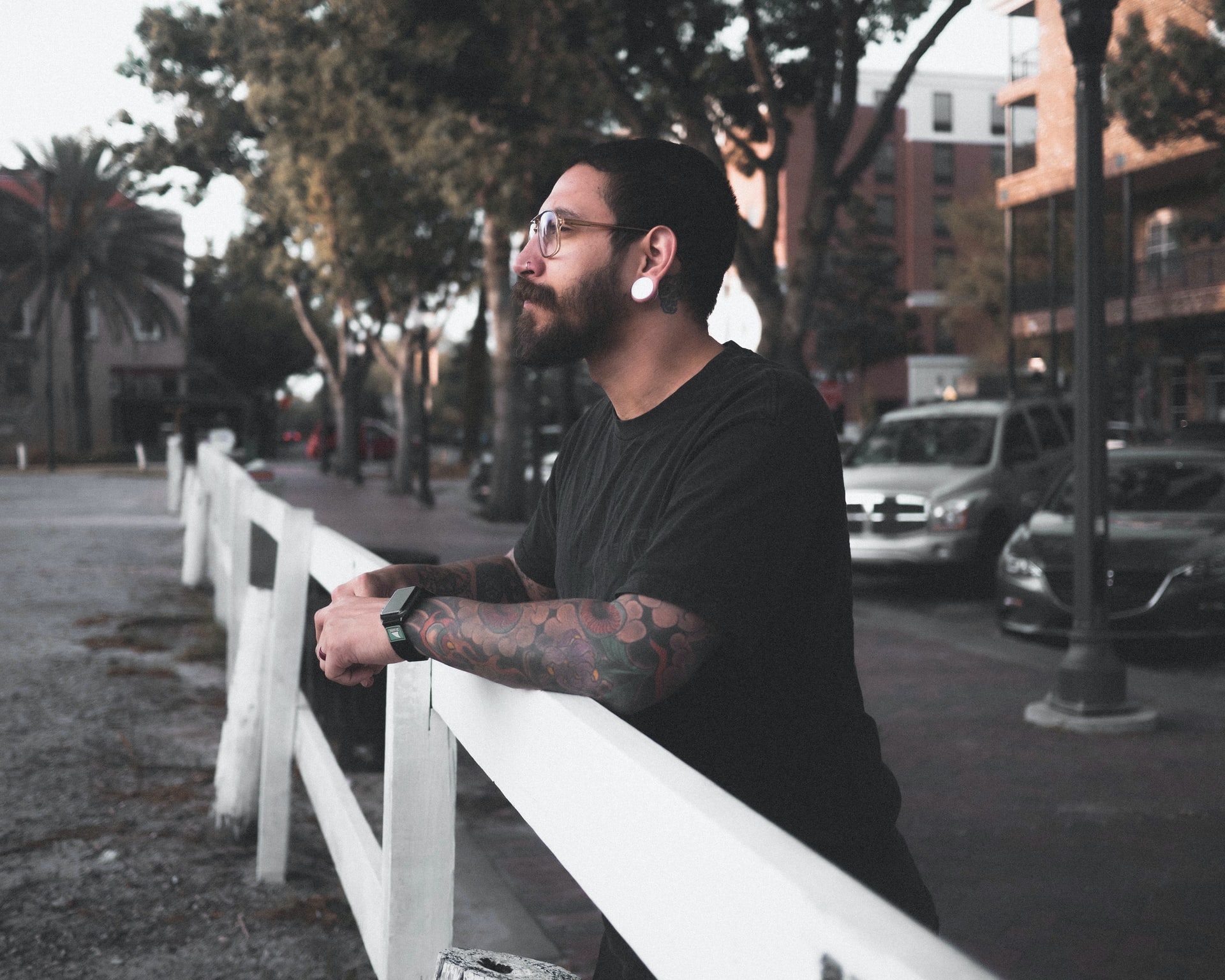 tattooed man with glasses and piercing leans on white rail during daytime