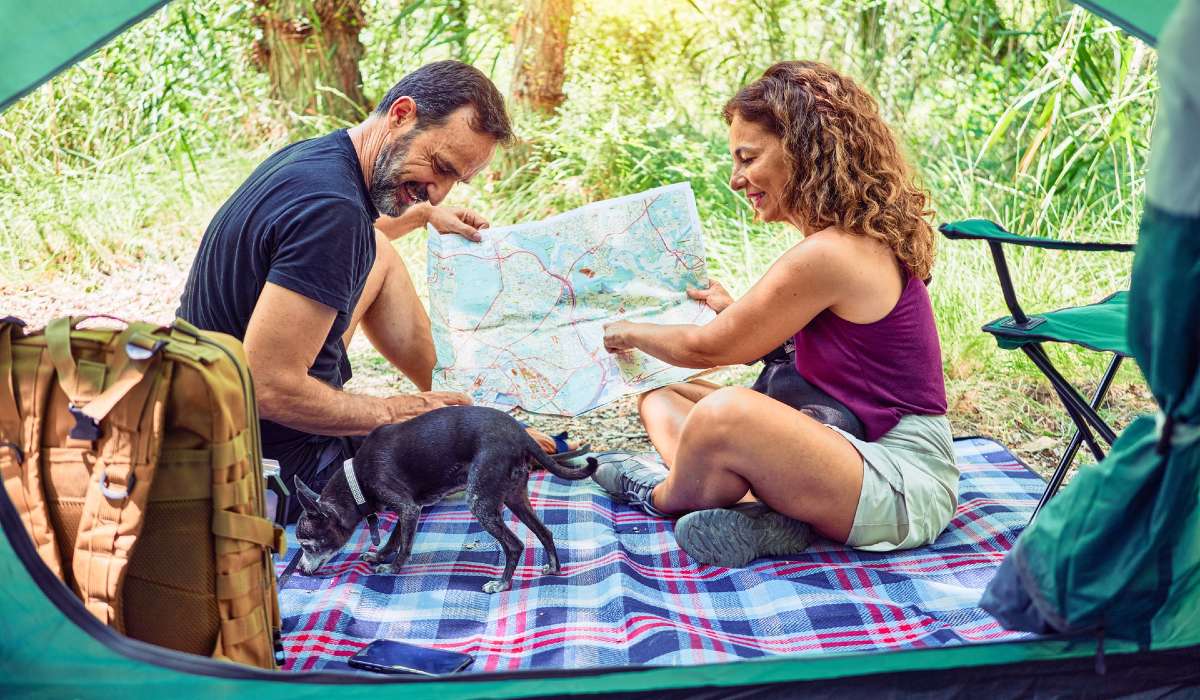Two people sitting on blanket out in nature, reading a map, little dog nearby, edges of a tent framing the picture