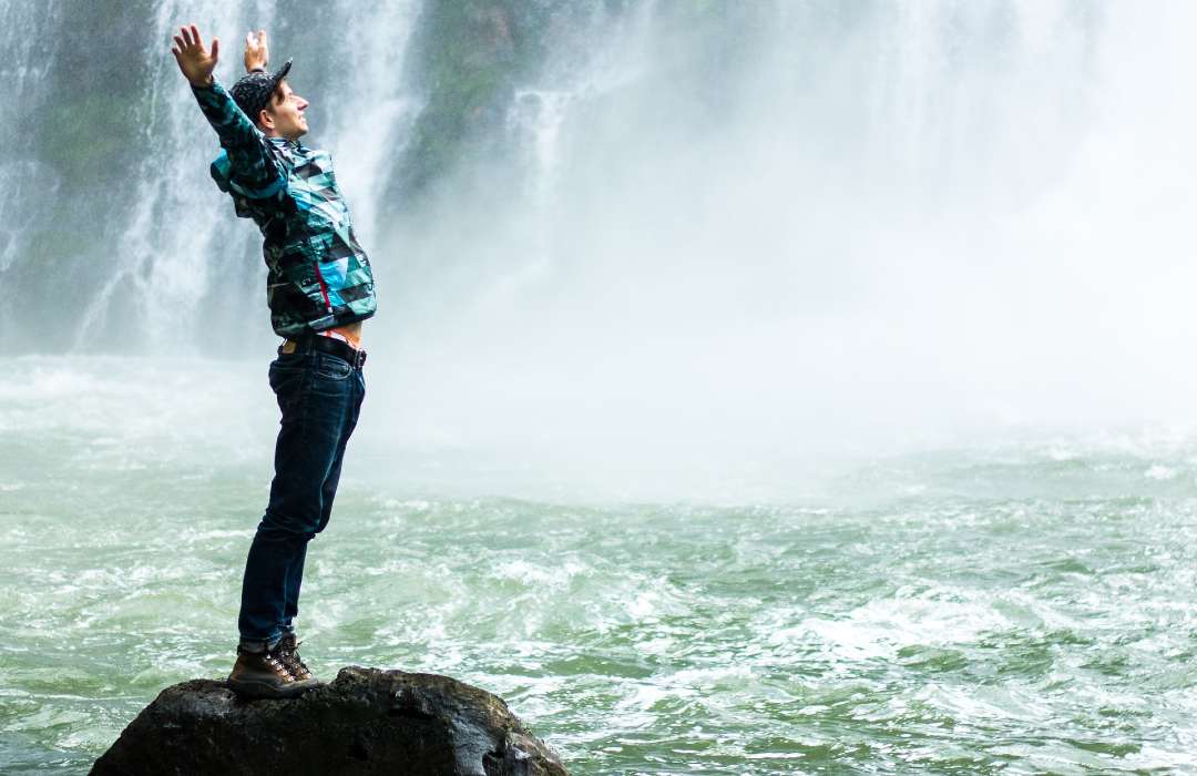 Person stands on rock in body of water next to waterfall, hands reaching skyward
