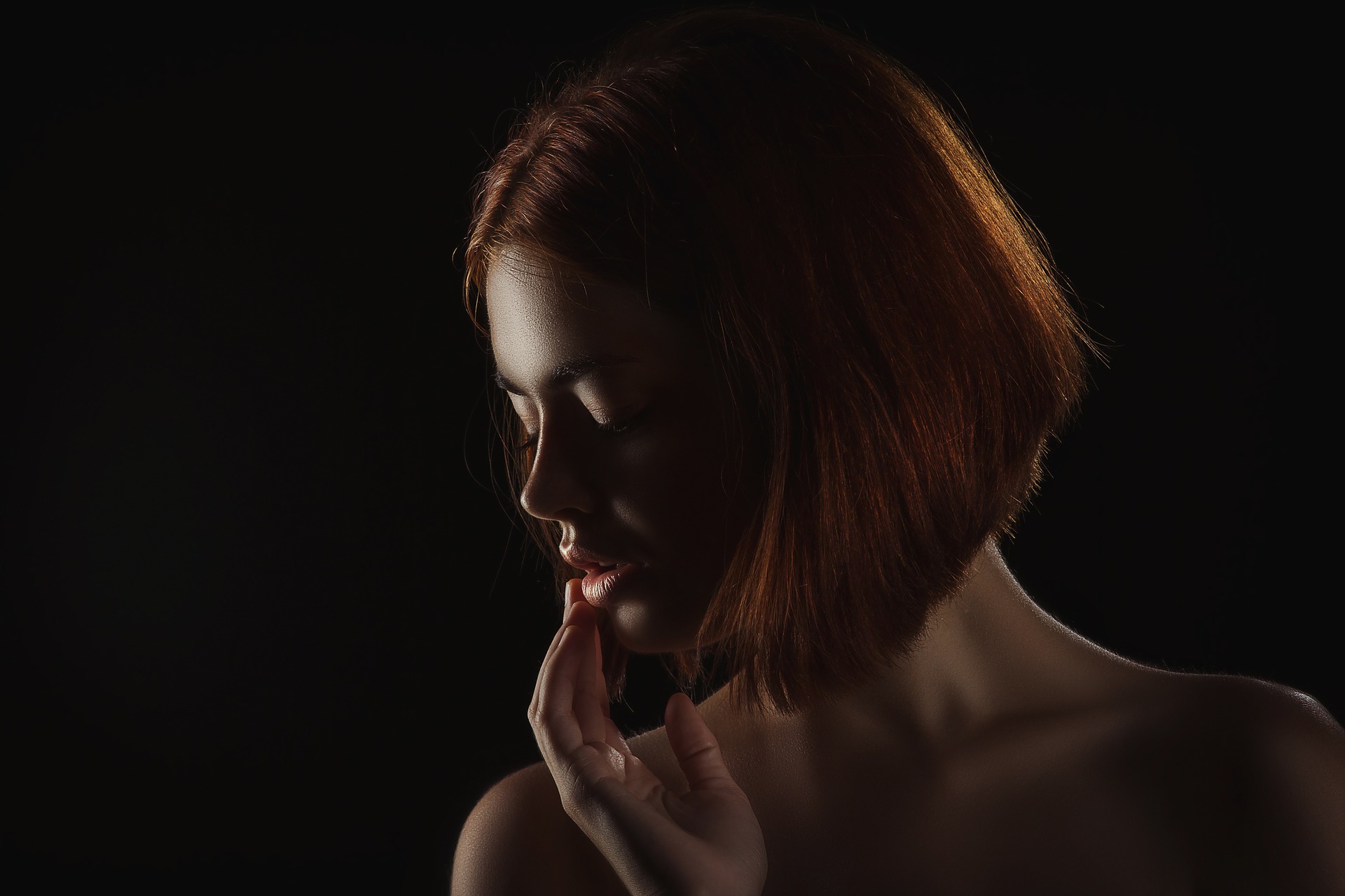 artistic shot of woman with red-haired bob-cut and no top on touching her lips, black background