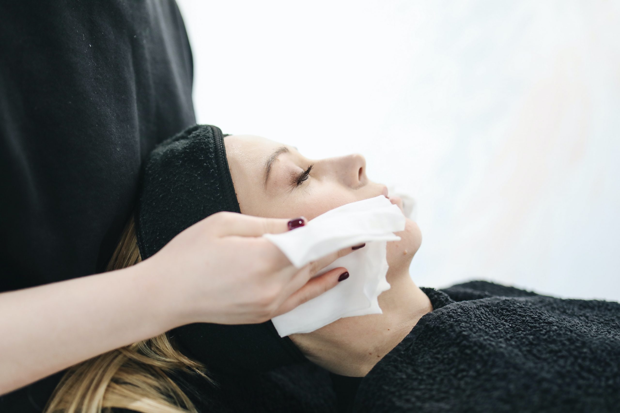 woman lying down, eyes closed, second person cleaning her face with cloth